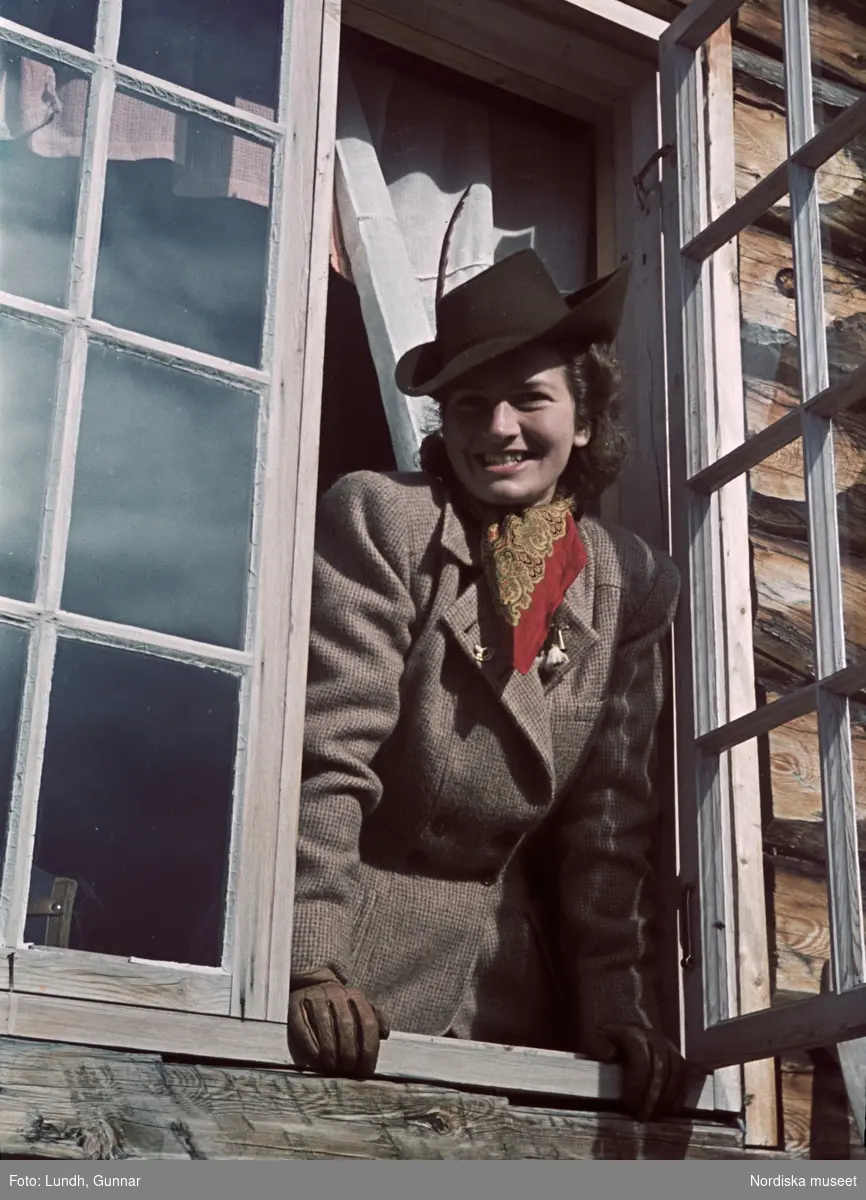 Tweedeklädd kvinna i hatt blickar ut genom ett öppet fönster.