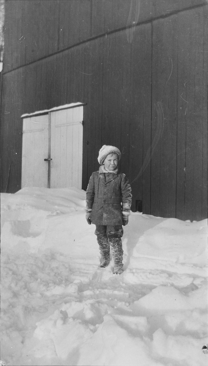 Iacob Ihlen Mathiesen som er fotografert foran en låve/driftsbygning. Guttenhar snø på klærne og snøen rundt ham er delevis måkt/opptråkket samt ren og fin. Det er vinter på Linderud Gård.
