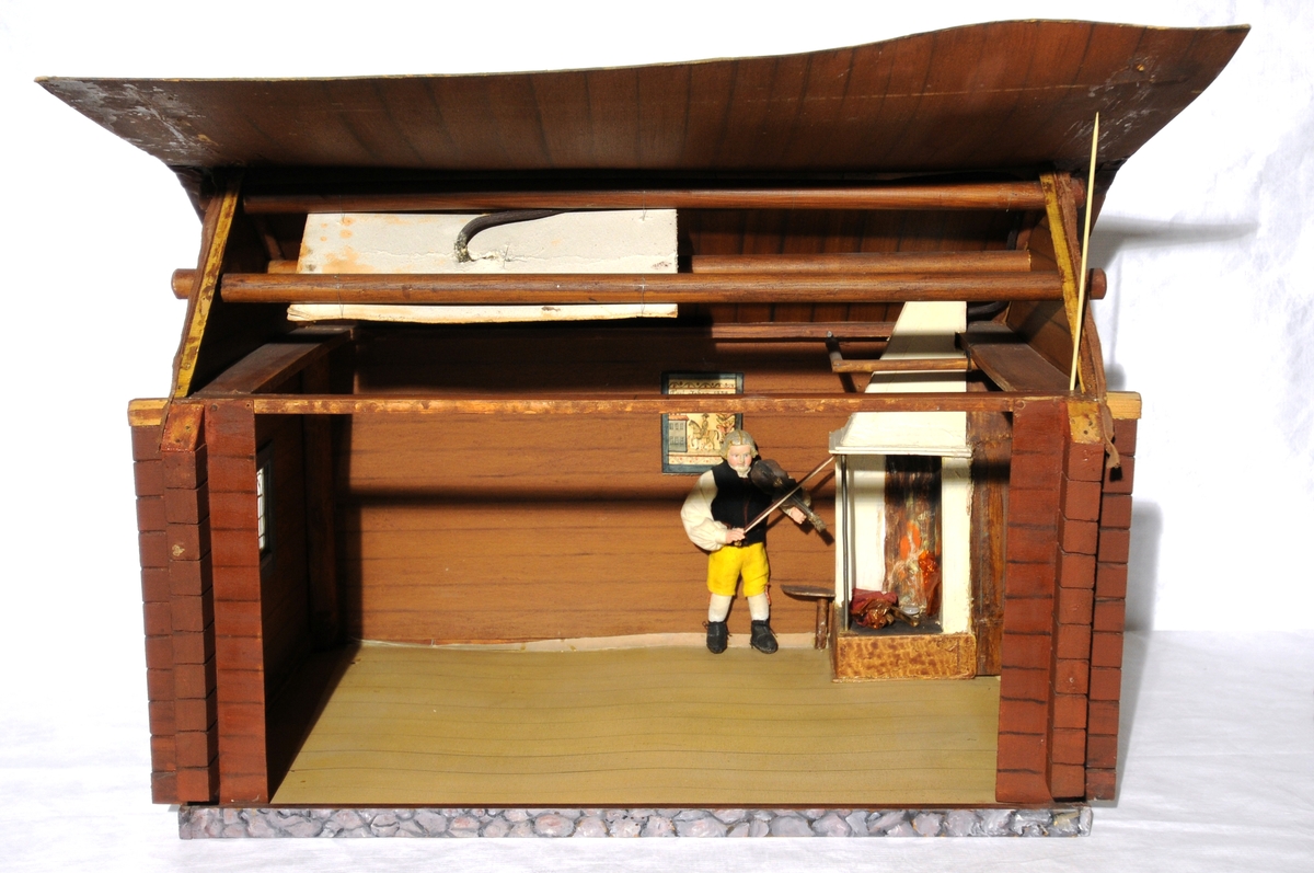 Dukkestue i form av svensk hytte med påmalte tømmerstokker, grunnmur og takstein i tegel. En vegg kan løftes bort og taket kan også åpnes. Stuen har tre stk vinduer med påmalte blysprosser. Inne i stuen er peis, bord og en dukke, en man i svensk folkedrakt med fele, fastmontert. På veggen henger et kurbitsdekorert maleri av Carl Johan.