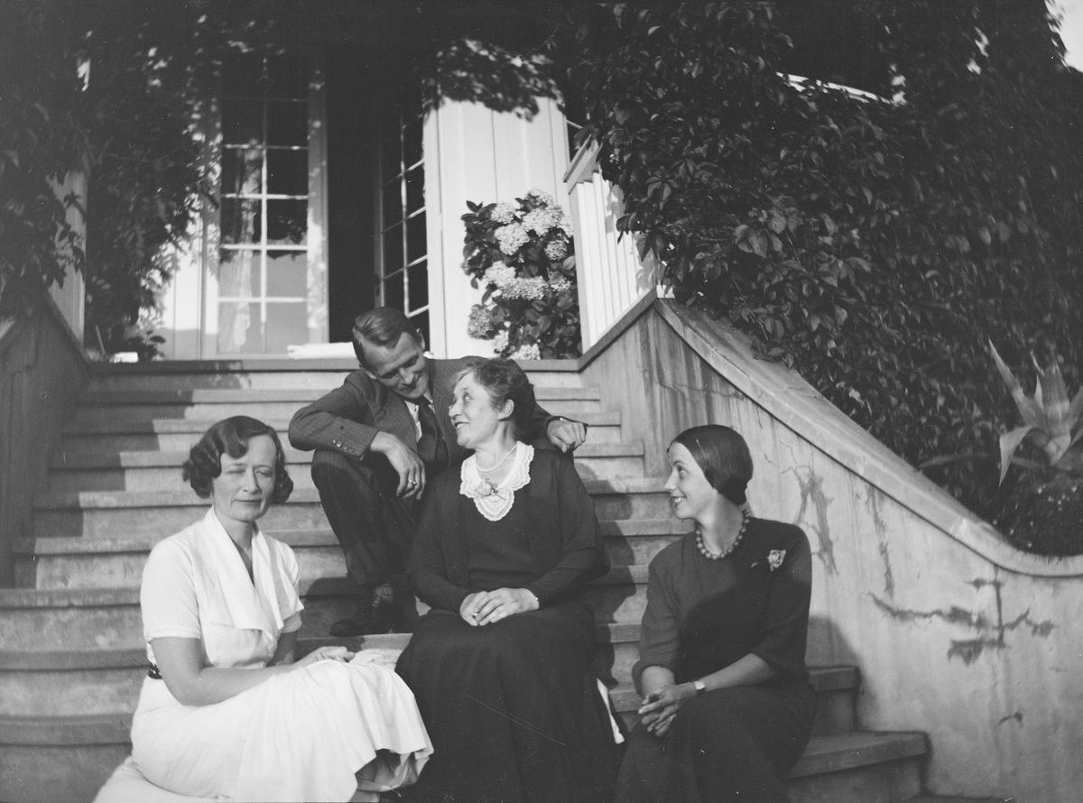 Tre kvinner og en mann sitter på trappen opp til verandaen fra den "Victorianske hagen" på Linderud Gård. De har tatt med puter som de sitter på. Verandaen er dekket av klatreplanter.