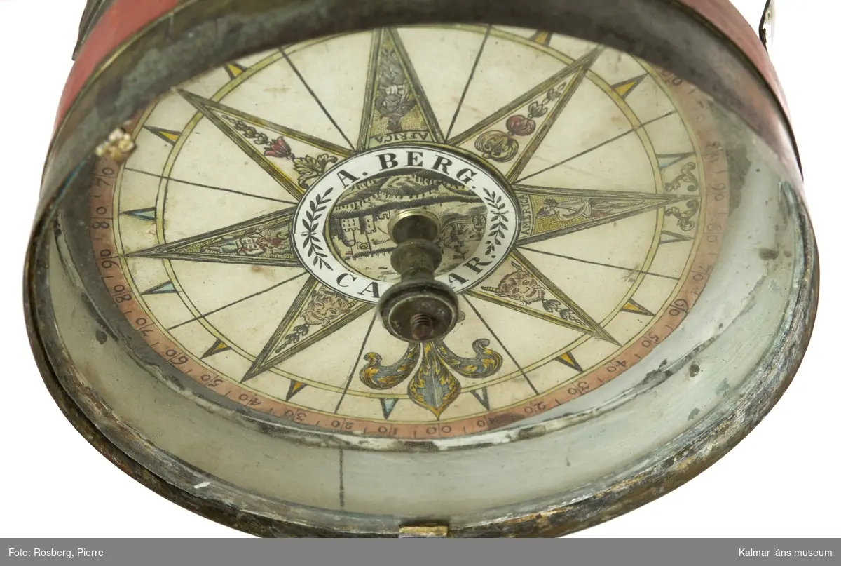 KLM 44862. Kompass, skeppskompass av metall, glas. Huset av metall översidan krönt av en kronliknade dekoration av metallband. Del av huset målat rött. En så kallad skvaller-kompass eller enligt Ekbohrn, Nautisk Ordbok, 1840: häng-kompass, hänger under skylight i kajutan sålunda att på den kan ses, äfven för kontroll, huru rorgängaren styr eller ligger an. Kompassen signerad, A. Berg. Calmar.
