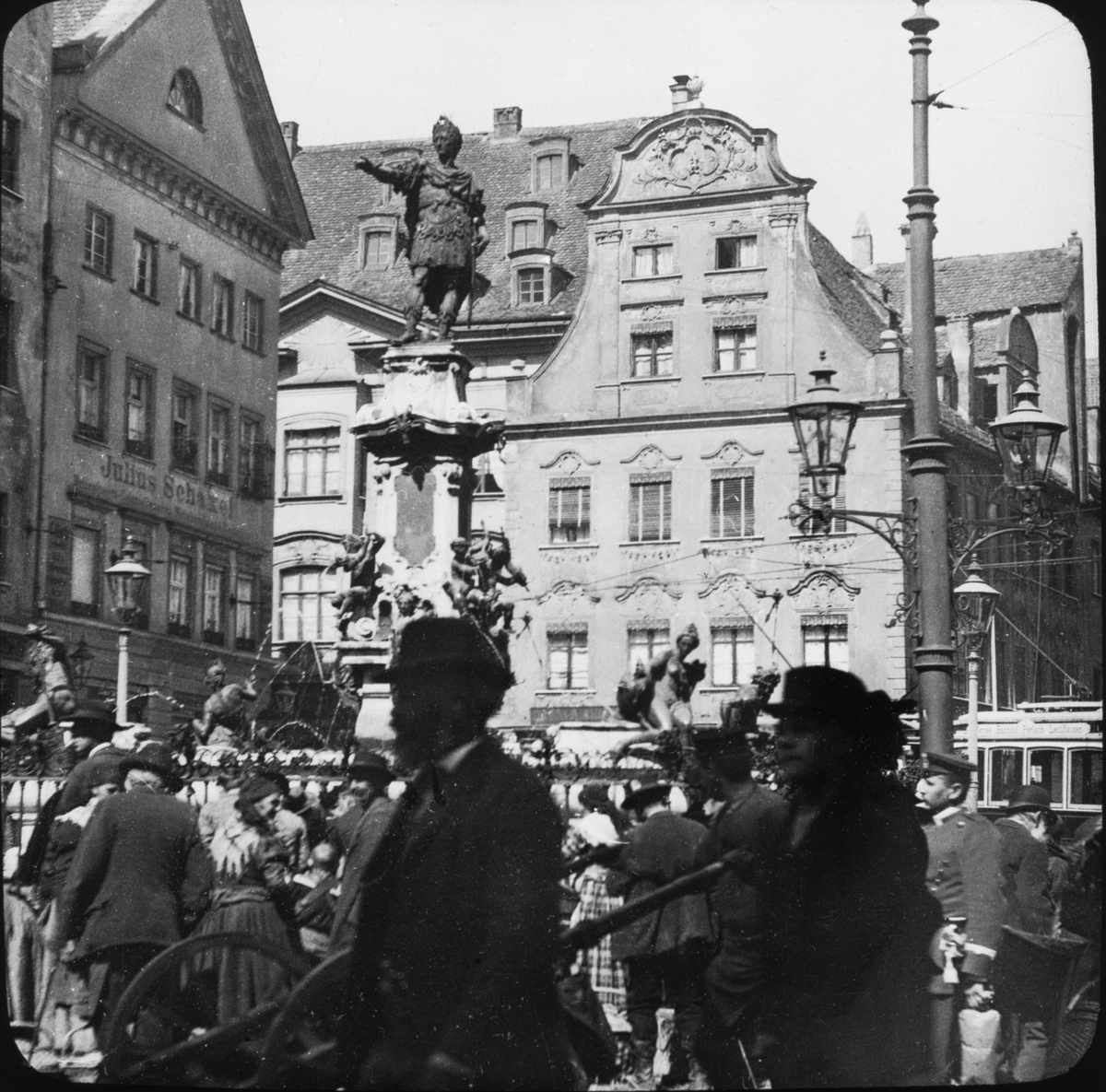 Skioptikonbild med motiv av folkvimmel kring Augustbrunnen, fontän i Augsburg.
Bilden har förvarats i kartong märkt: Augsburg 1901. 13. Text på bild: "Wiirzburg 1901".