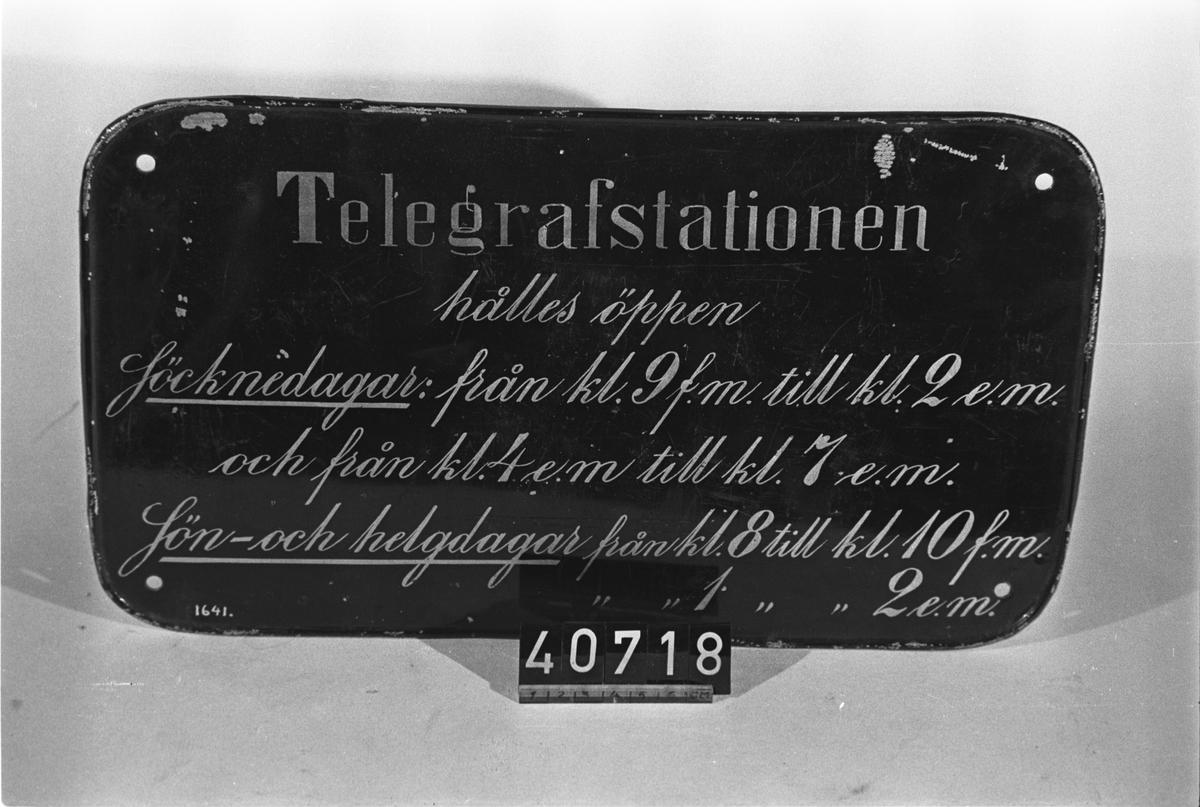 Stationsskylt i svartlackerad plåt, typ "Nr 3". Text: "Telegrafstationen hålles öppen Söcknedagar: från kl. 9 f.m. till kl. 2 e.m. och från kl. 4 e.m. till kl. 7 e.m. Sön- och helgdagar från kl. 8 till kl. 10 f.m. från kl. 1 till kl. 2 e.m."