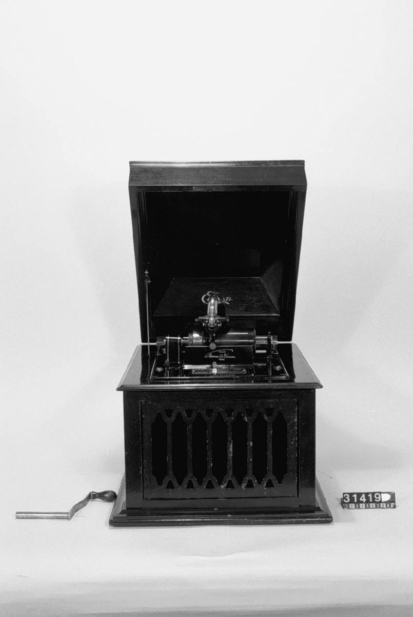 Fonograf inbyggd i låda av mahogny med uppfällbart lock, med tratt direkt från ljudarmen och utmynnande i lådans framsida. Gallret vid trattens mynning sitter löst