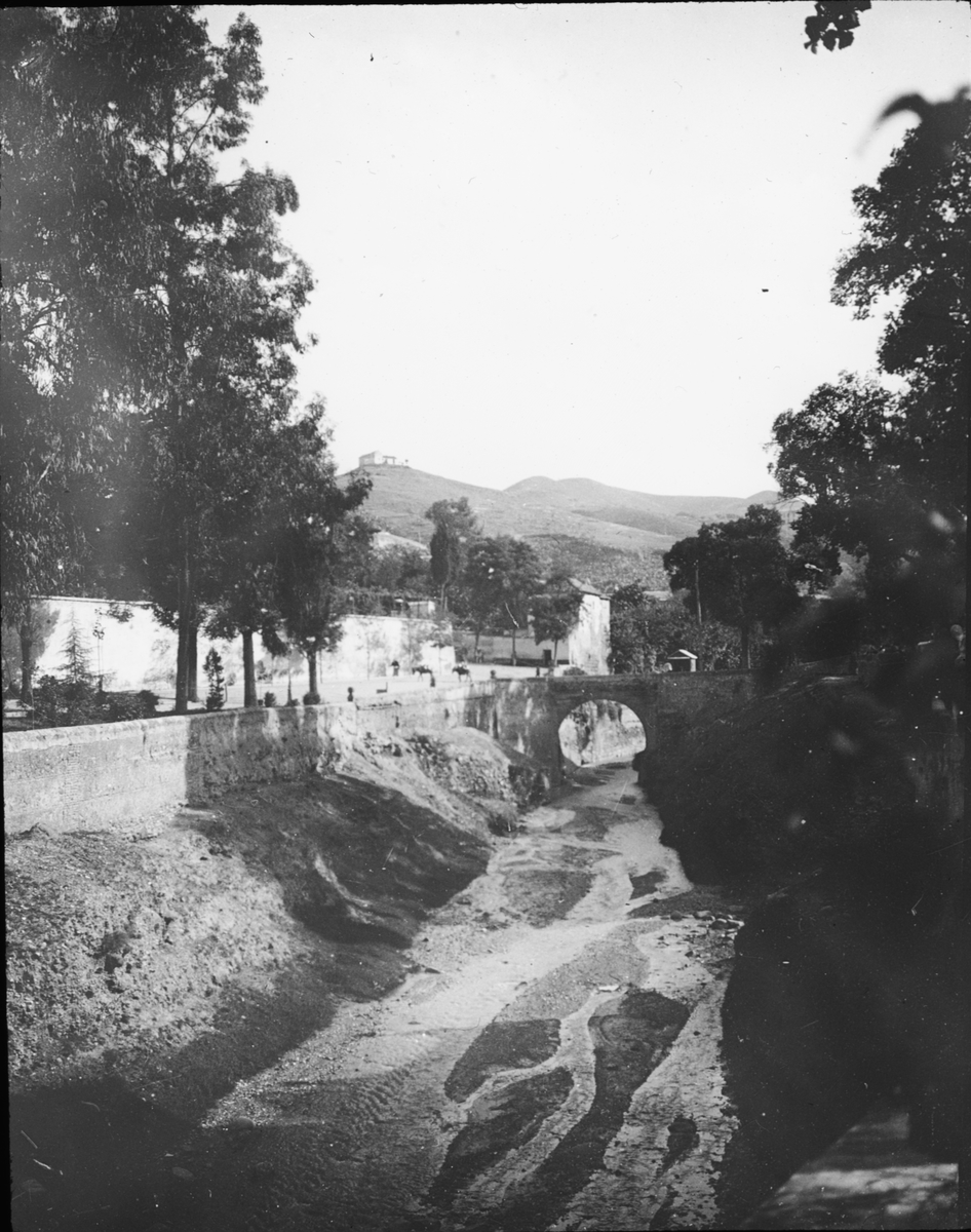 Skioptikonbild med motiv av uttorkat vattendrag, Darro, i Granada.
Bilden har förvarats i kartong märkt: Höstresan 1910. Granada 9. N: 29. Text på bild: "Darro vid vatten".