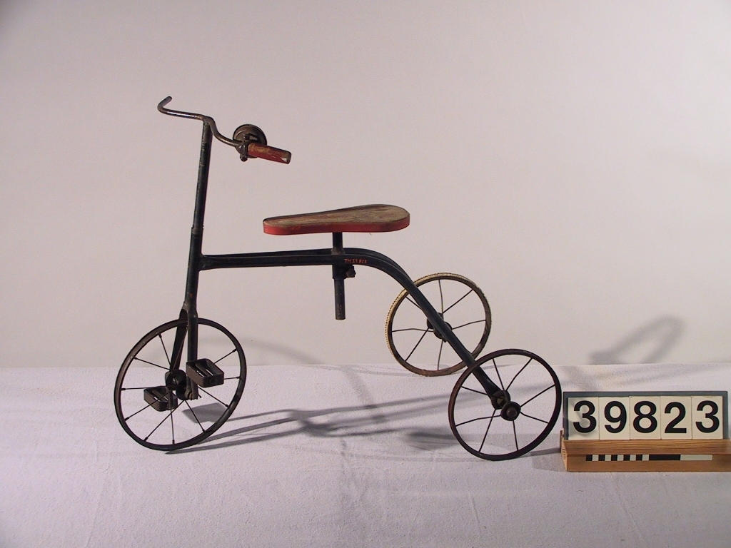 Barncykel, trehjuling med röd träsits. Cykeln har blivit utrustad med en ringklocka. Ett handtag saknas liksom däcken.