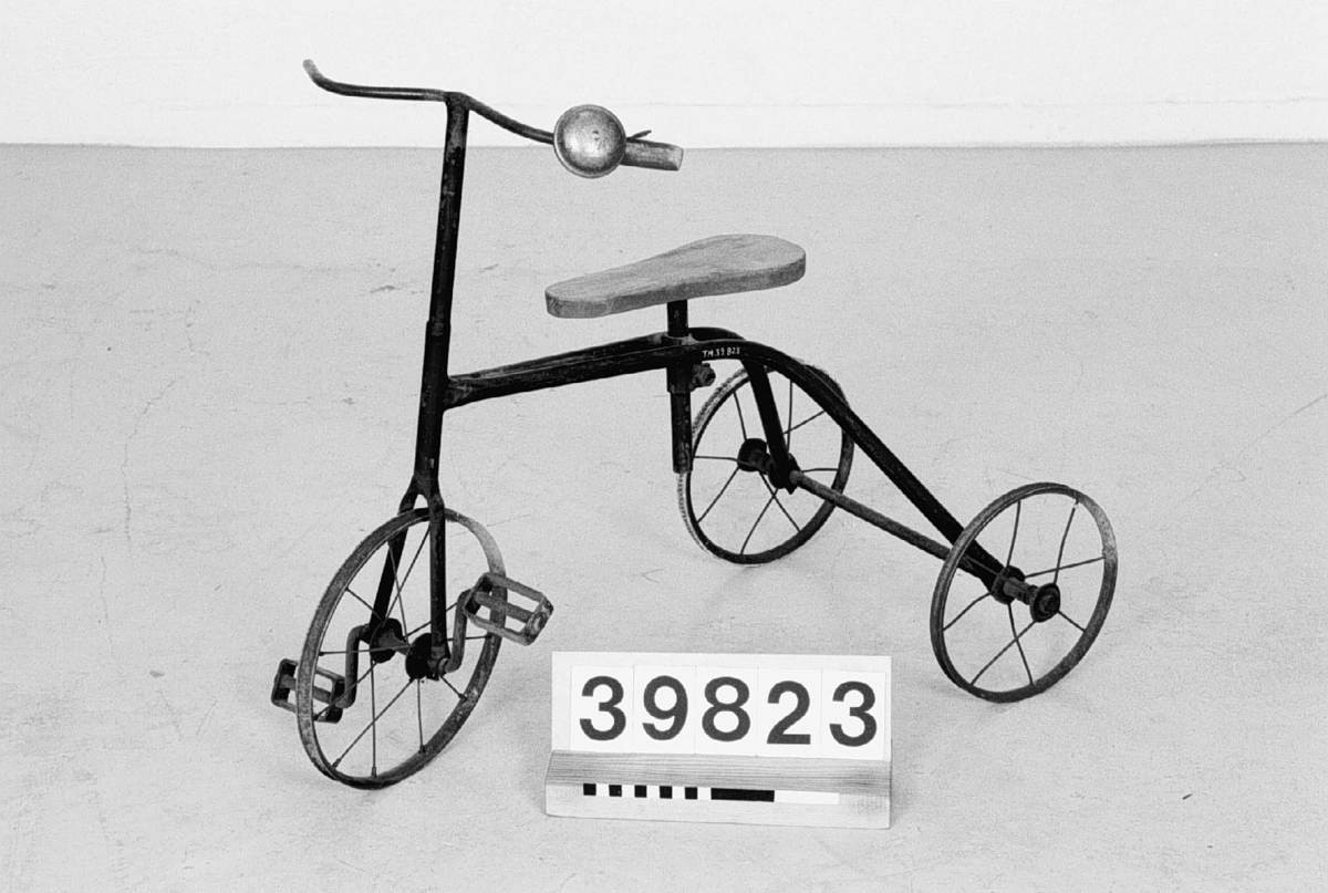 Barncykel, trehjuling med röd träsits. Cykeln har blivit utrustad med en ringklocka. Ett handtag saknas liksom däcken.