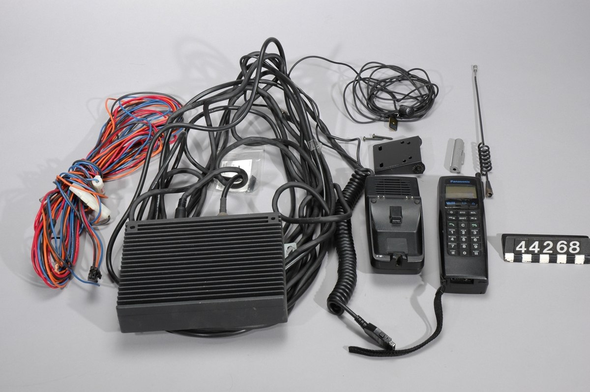Panasonic NMT mobiltelefon typ EB-3710A med utdragbar antenn och handlovsrem. Batteriet ej original, daterat 97-02. Tillhörande signalförstärkare "Booster/charger" typ EB-PO515 och handsfreenhet "Handsfree cradle" typ EB-JO514. Yttre antenn för bil finns, men utan antennfot och kabel.