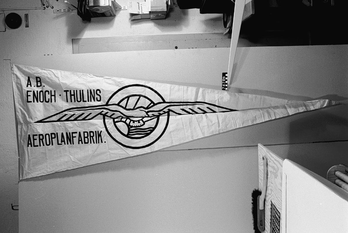 Av vitt bomullstyg med reklamtext i svart för AB Enoch Thulins Aeroplanfabrik, Landskrona, använd vid provflygningar. Text i svart färg: "A.B./Enoch Thulins Aeroplanfabrik" och däröver stiliserad mås (fabriksmärke).