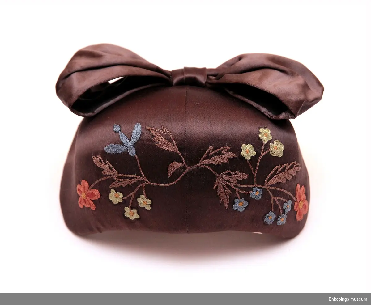 Bindmössa, en liten hätta/mössa för kvinnor. Den är brun med blommönster på och rosett på baksidan.