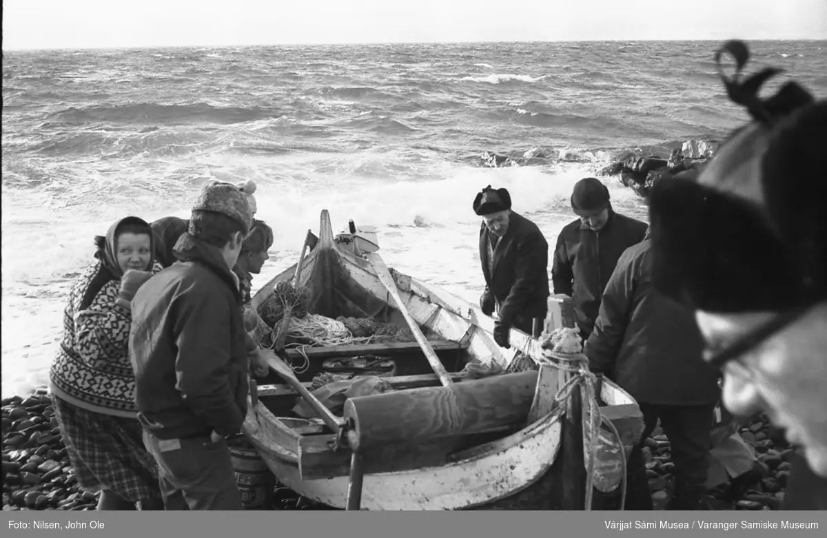 Brødrene Andersen fått hjelp til å dra båten på land. Bildet er tatt i Gurluovttgohppi / Godluktbukt. Den unge mannen nærmest til venstre er en Johansen fra Mortensnes og kvinnen med ansiktet hitover er Johanne Pettersen. 28. februar 1967.