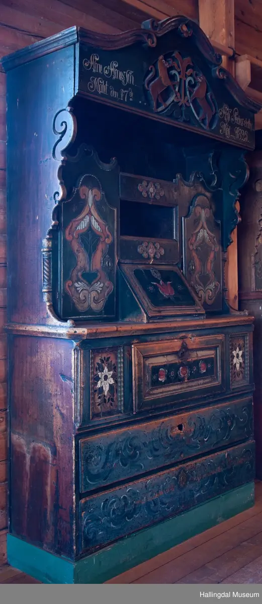 Delene satt sammen v.hj.a. tapping og trenagler.  Gavlen buede sider med skåret kronlist, delt i 3 profilerte, med opprullede ender.  Kronlister fortsetter på sidestykkene.  I toppen av kronlist hjerteformet, gj.brutt utskjæring opp ned.  Gavlens sentralfelt utskjæring i dyp flatskurd.  To løver, vendt mot hverandre, griper om hellebordskaft.  En stake som øverst kløyves i to bjørnehoder i profil skiller løvene.  Gavlen (el. baldakinens) forplate danner fremsiden i skuff i skapets bredde og dybde.  Overskapet:  Åpen hylle øverst, ca. 25 cm høy.  Under hyllen er det delt i 3 felt.  Ett skap i hvert sidefelt.  Midtfeltet har 2 skuffer adskilt av åpen hylle.  H. Skuffe: 12,5 cm.  H. åpent rom: 17 cm.  Under nederste skuff, skap med skråsider som stikker ca. 15 cm ut fra forveggen på overdelen.  Skapet dekkes av liggende skråklaff.  Festet jrenbeslag og hengsler nederst.  
En stor skuff i hele skapets bredde, 6 cm høy nederst, 32 cm dyp.  Over skuffen er rommet delt i 3 felt.  Hvert sidefelt har 2 skuffer og midfeltet en dyp skuff.  Midskuff har fylling med utskåret medaljong, klaff og hylle og skuffen rødmalt. .................................................