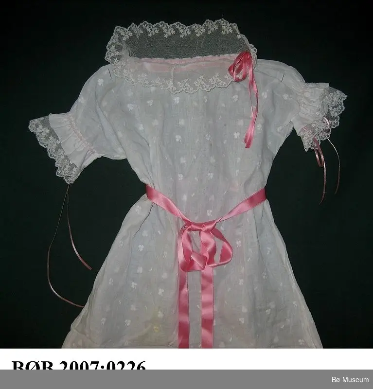Kvit dåpskjole med bloder og rosa band. Kjolen er sydd av eit tøystykke. Det er klypt hol til hovudet og kjolen er sydd saman i sidene med fransk saum. Kjolen blir heldt saman i livet med eit rosa silkeband.