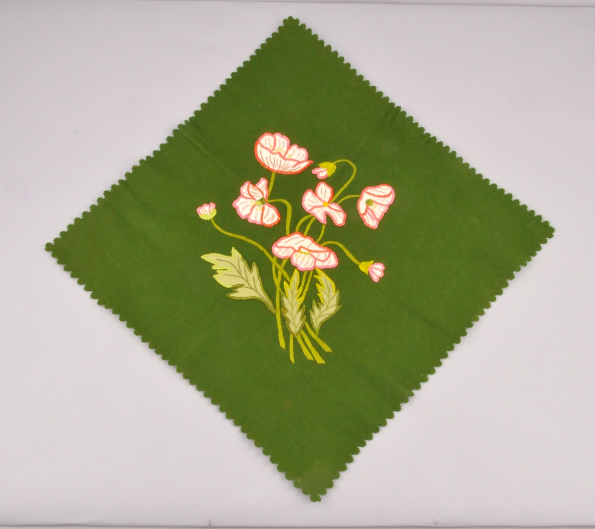 Grøn kvadratisk lita brikke av klede med rosa og kvitt broderi med blomstermotiv. I applikasjon og silkebroderi. Klipte  tunger rundt kantane.