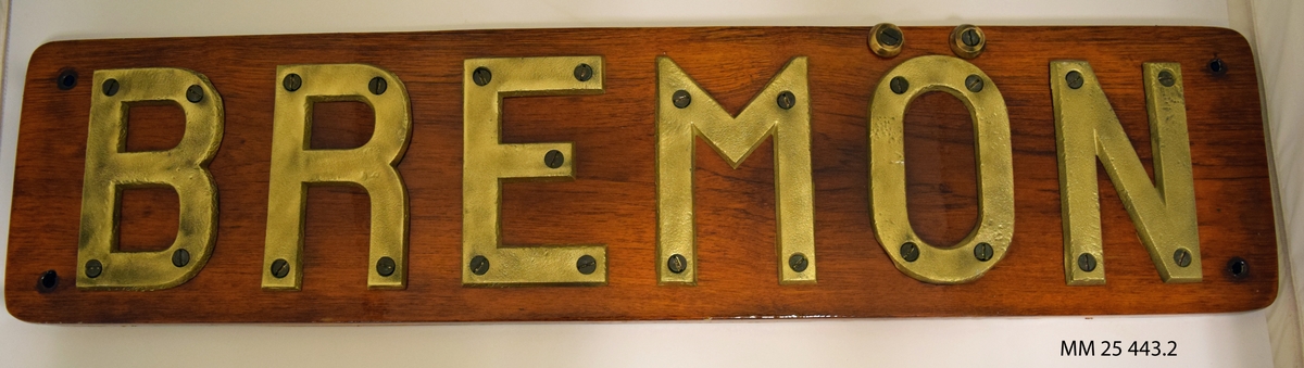 Namnbräda. En rektangulär träplatta, fernissad, med bokstäver i stål skruvade i plattan bildar namnet "BREMÖN".