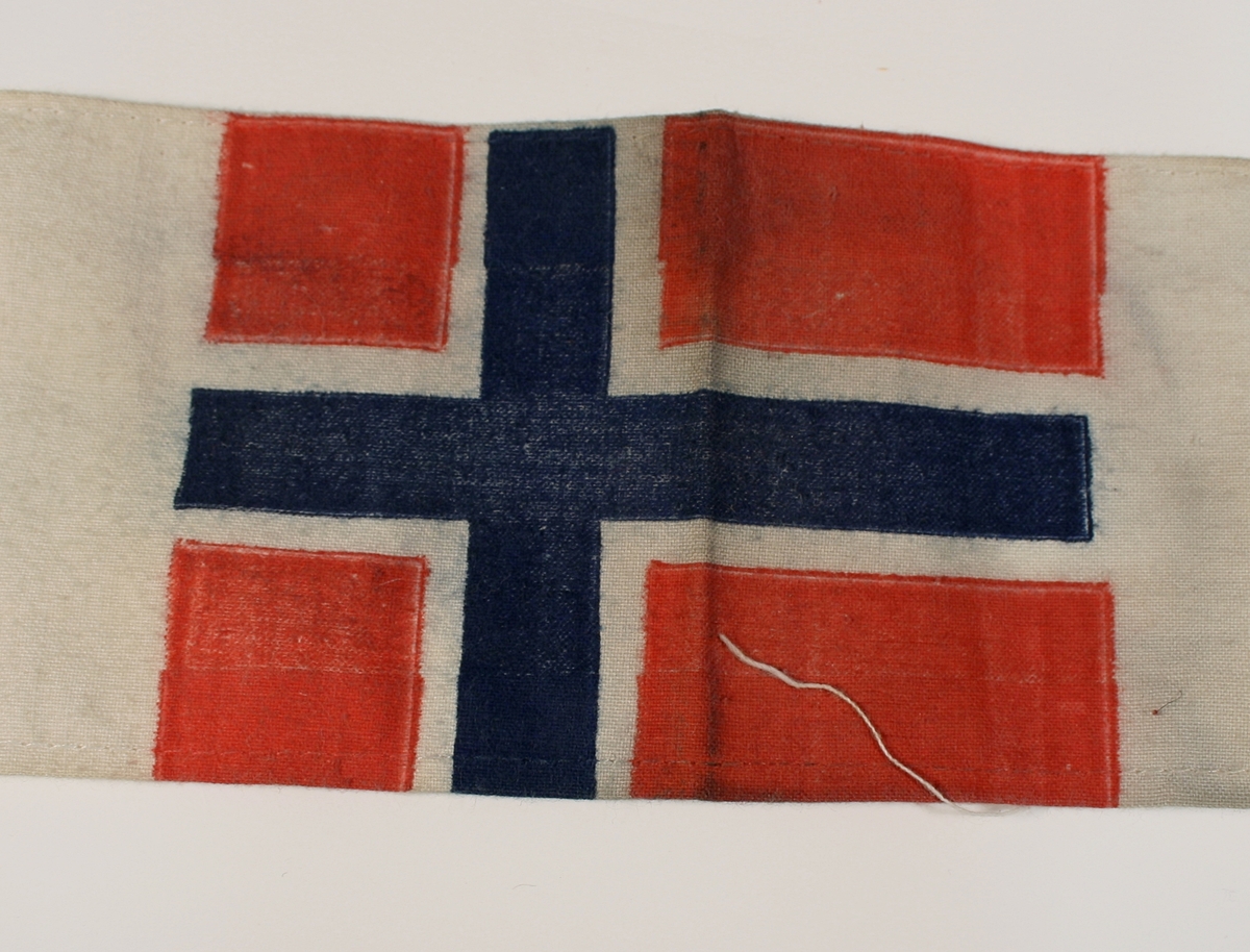 Armbind i hvitt stoff med vit bunn med det norske flagget.