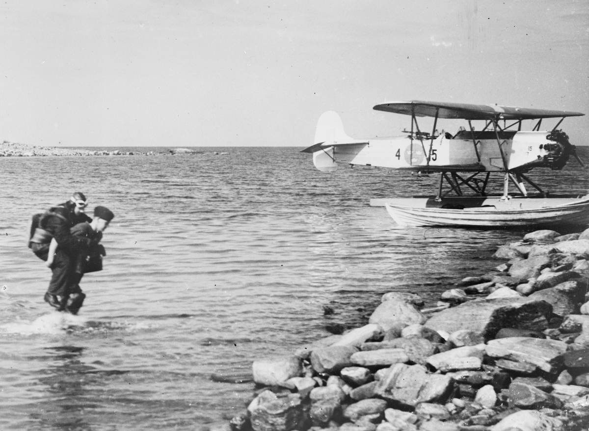 En militär flygare bärs i land av markpersonal. I bakgrunden är ett flygplan S 6H märkt nummer 15 från F 4 Frösön förtöjd vid stranden.