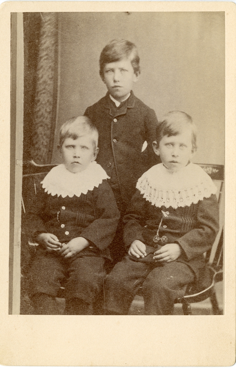 Portrett av tre gutter. Alle er kledd i mørke knebukser og jakker. De to minste har store hvite heklete krager.