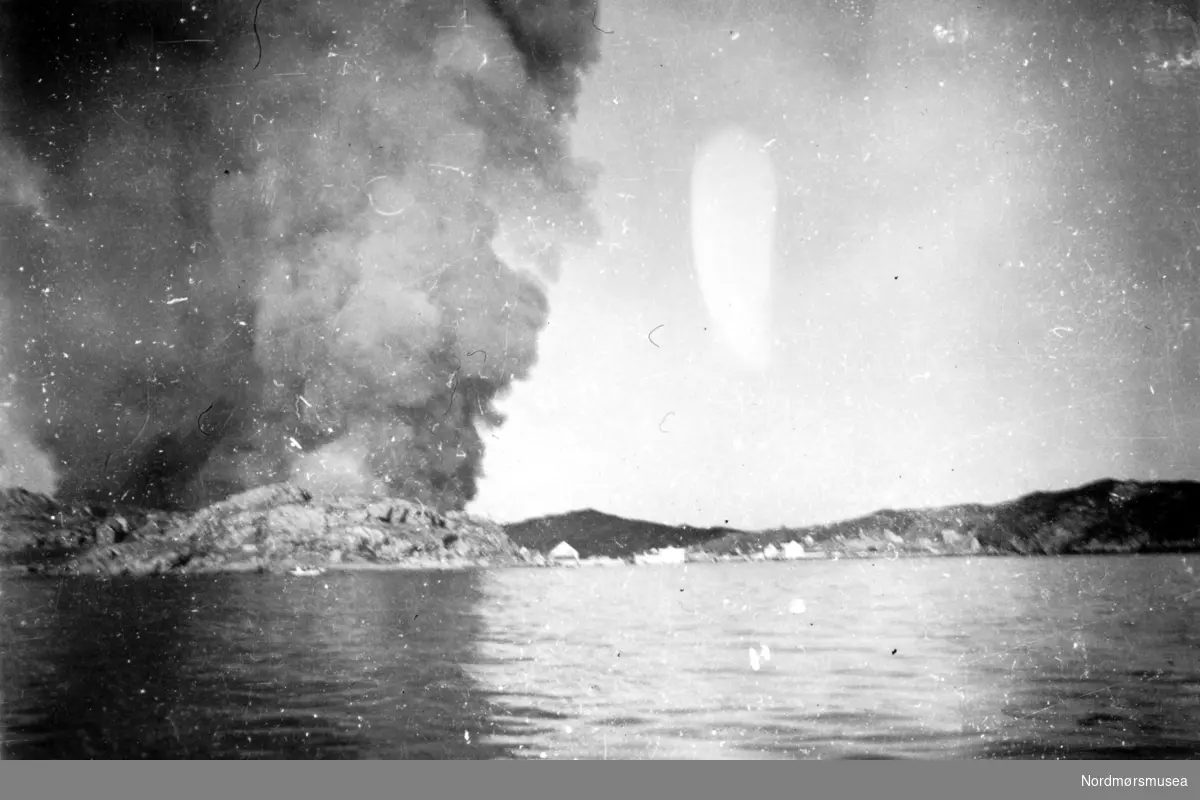 Andre verdenskrig har kommet til Kristiansund, hvor vi ser vestre bydel i brann. Bildet er datert 28. april 1940, som er samme dag som byen ble bombet for første gang, og fotografert fra Bremsnesfjorden.  Fra Nordmøre museums fotosamlinger.