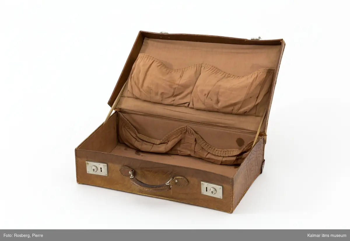 KLM 28668:243. Väska. Liten rektangulär resväska av ljusbrunt läder med handtag och två förnicklade lås. Två nycklar finns. Skoningar av hård papp på två av bottenhörnen. Foder av ljusbrun moiré med fickor i botten och lock. Måtten tagna med väskan i liggande position.