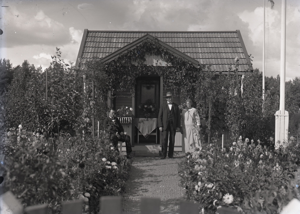 En stuga, två män och en kvinna i Kalmar södra koloniområde, fotograferat omkring 1930. Kalmar södra koloniförening grundades 1917 och har idag 105 kolonilotter. Området ligger strax söder om länssjukhuset i Kalmar med huvudingång från Stensbergsvägen.