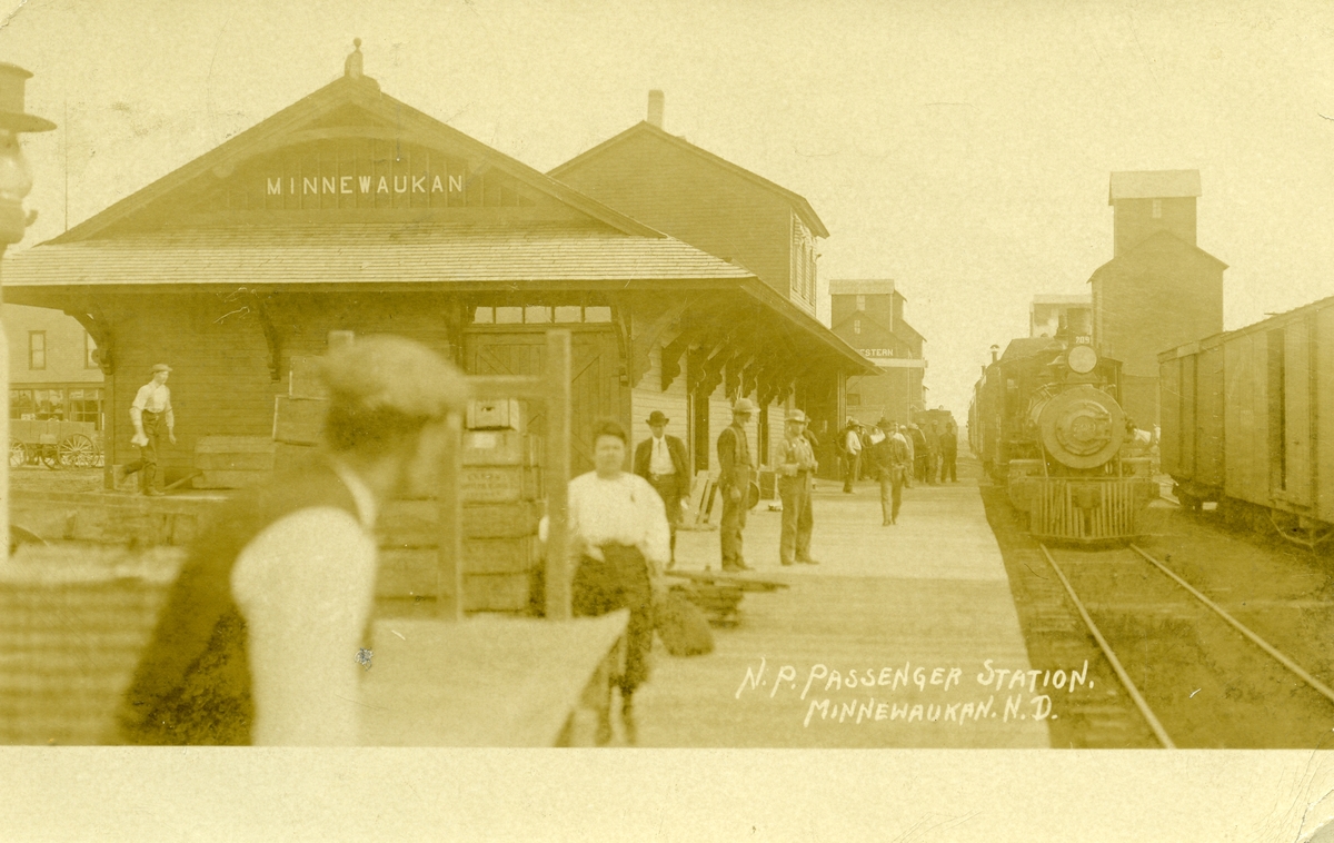 Postkort med motiv fra Minnewaukan passenger station. Bildet viser stasjonsbygningen, toget som står på stasjonen og flere passasjerer/personer på perrongen.