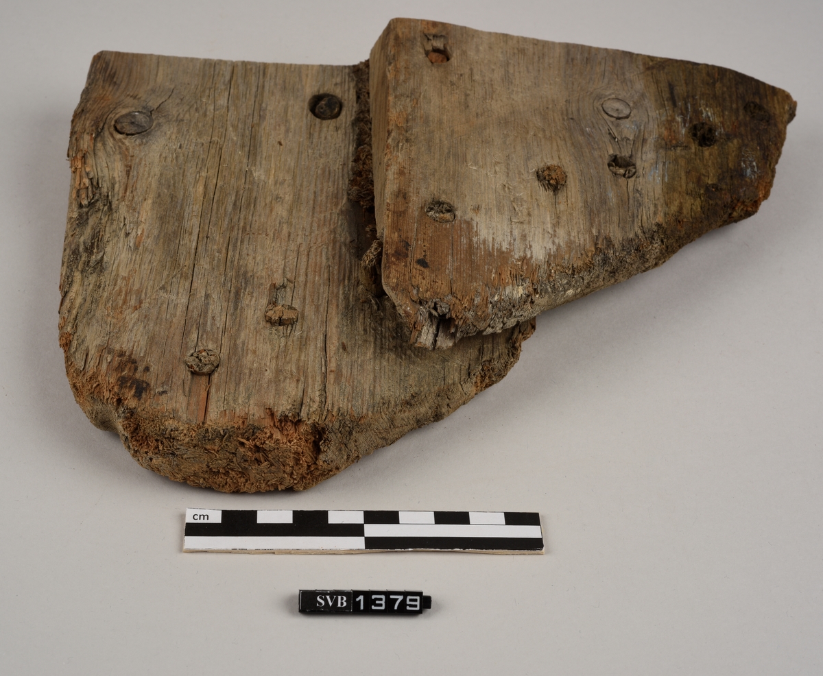 Del av et båtbord. Delen ble saget av. Det er hull til vidje og plugger. Delvis er det rester av både vidje og plugger. Treet fremstår ubehandlet. Det er et materiale som ligner torv/mose mellom bordene.