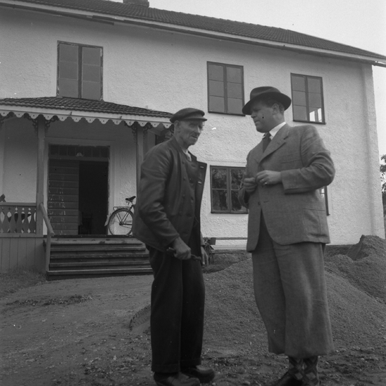 Foto av två män - den ene klädd i kostym,hatt och den andre klädd i jacka, byxa och en skärmmössa.
I bakgrunden skymtar ett bostadshus med en cykel på verandan.