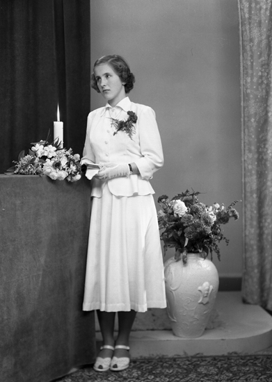 Foto av en ung kvinna i vit, tvådelad konfirmationsklänning.
Hon håller en liten psalmbok i händerna.
Helfigur. Ateljéfoto.