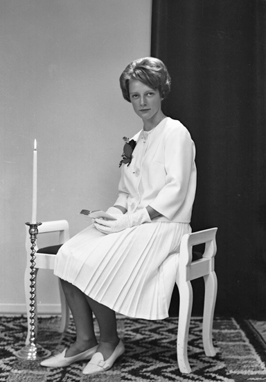Foto av en kvinnlig konfirmand i vit konfirmationsklänning. Hon sitter på en vitmålad, stoppad bänk med en golvljusstake vid sidan.