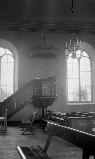 Foto på en predikstol med kyrkbänkar längst fram i bild, det hänger en ljuskrona i taket.