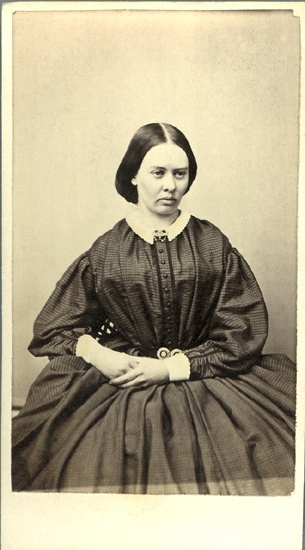 Porträtt (knäbild, halvprofil) av en sittande kvinna i mörk klänning med vit spetskantad krage. Vid kragen skymtar en brosch.