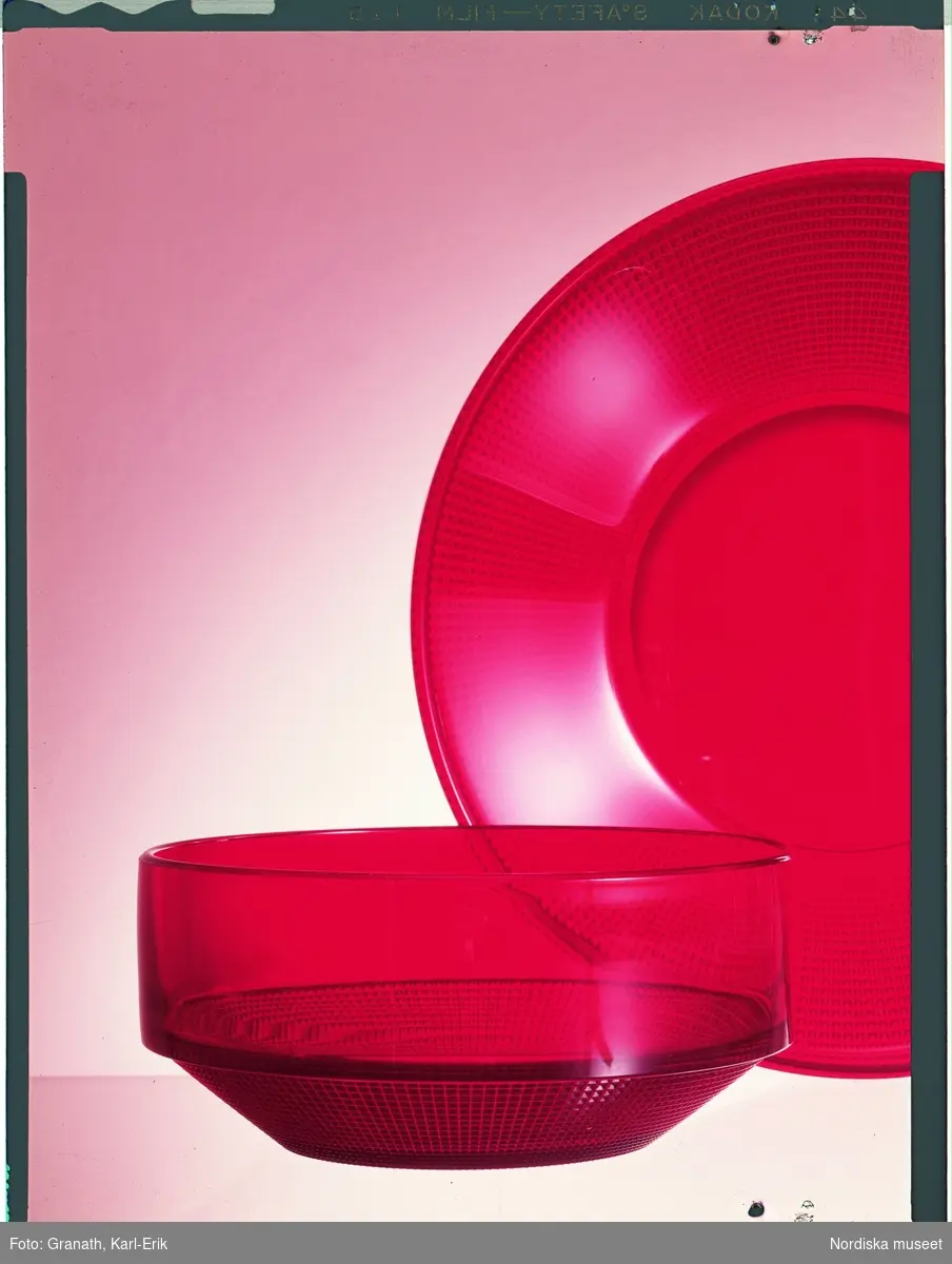 Röd design. Stilleben av ett upprättstående fat och en skål, båda av rött, transparent glas med ett rutnätsmönster.