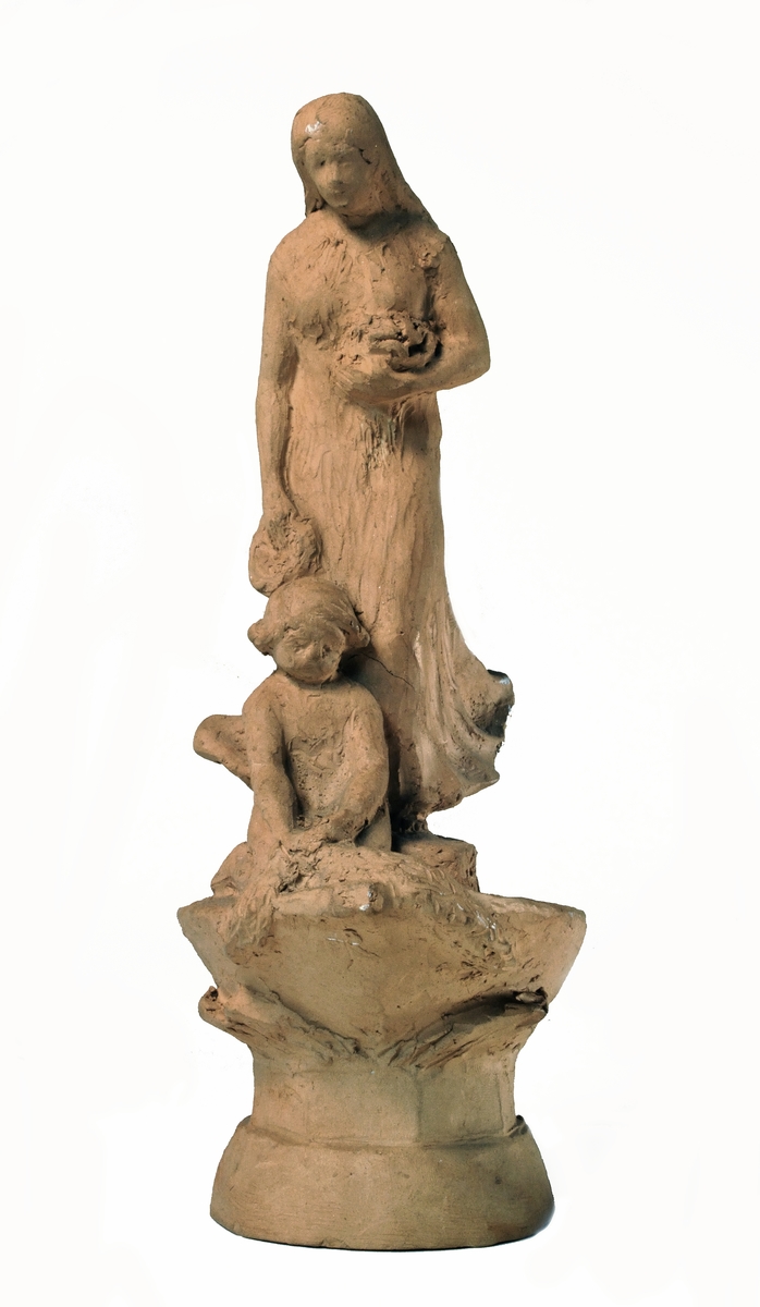 Skulpturskiss av Ida Matton till gravmonument för redarfamiljen Brodin i Gävle. Kvinna med blommor i händerna med ett barn med blomgirlang sittande framför fötterna. Båda placerade på en trekantig sockel som liknar stäven på en båt.