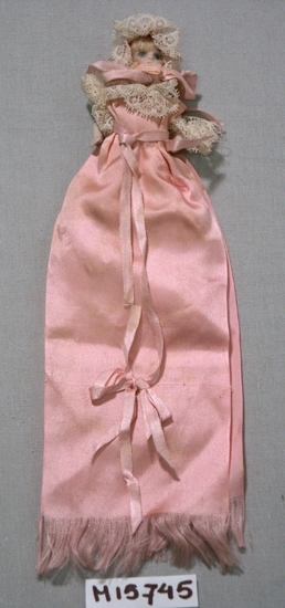 Docka, liten ca 100 mm lång, av porslin, med blont hår.
Dockan har en klänning gjord av ett skärt sidenband. Spets kring hals och ärmar.
På huvudet en skär sidenhätta med spets.
Dockan har en underklänning tillverkad av vit muslin, uddig i kanten och kråksparkar 
sydda med skärt silke.

Inskrivet i huvudbok 1954.