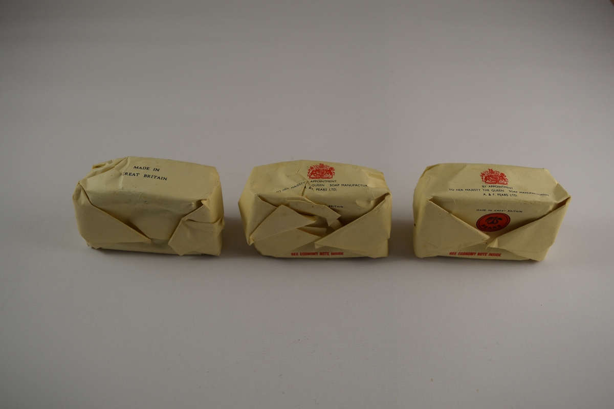 Tre såpestykker innpakket i papiremballasje med sort og rød skrift. Ovale såper brunt, gjennomsiktig materiale