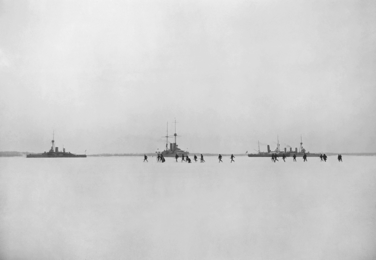 Svensk militär under språngmarsch på isen framför fartyg i den svenska eskadern under Ålandsexpeditionen 1918. I bakgrunden ses från v till h pansarskeppen THOR och SVERIGE, isbrytaren ISBRYTAREN II samt pansarskeppet OSCAR II.