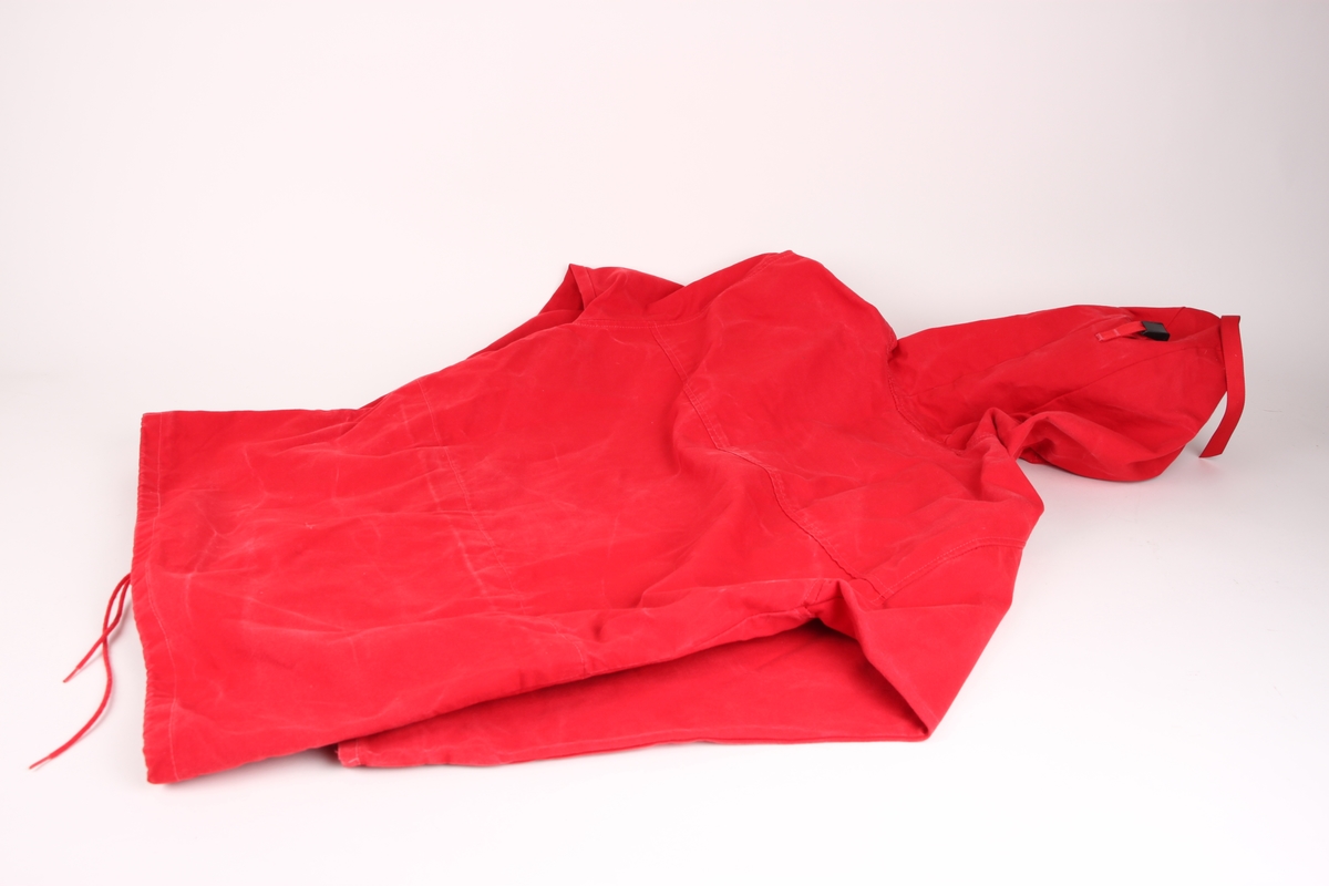 En rød vindanorakk i tykk bomull med glidelås og fire lommer med klaffer foran på jakken. Vindanorakken har også snøring i livet, samt stormhette med brem.