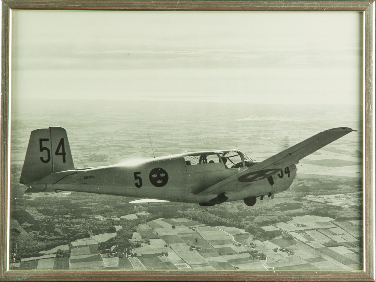 Flygplan SK 50 märkt nummer 54 från F 5 Krigsflygskolan i luften, flygande över landskap. Inramat foto.