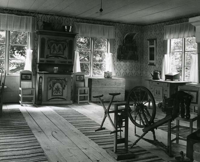 Interiör med schablonmålade väggar från Ersk-Anders gårdsmuseum i Skästra, Järvsö. De breda golvtiljorna av trä pryds med långa trasmattor och där står en spinnrock.  Det står ett stort och fint dekorerat skåp mellan fönstren.