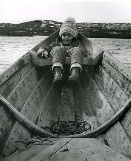 I fören på en träbåt sitter en liten flicka hopkurad. I fonden syns låga fjäll, Lappland.