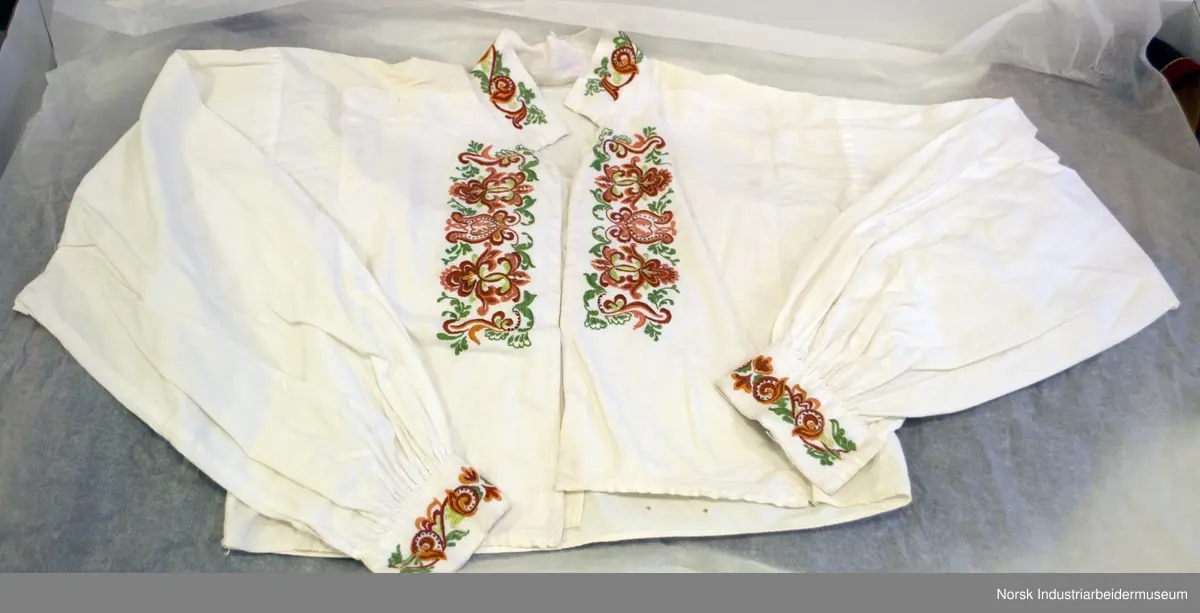 Plattsøm på kraven, mansjettene og bringa. Skjorten er åpen foran og helt ned.