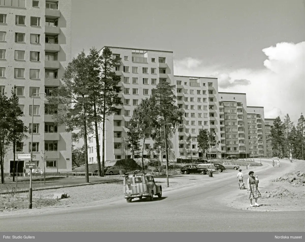 Vy över hyreshus i hörnet Glanshammarsgatan, Skyllbergsgatan i stockholmsförorten Hagsätra. Promenerande kvinnor och barn. En Volvo Duett i förgrunden.