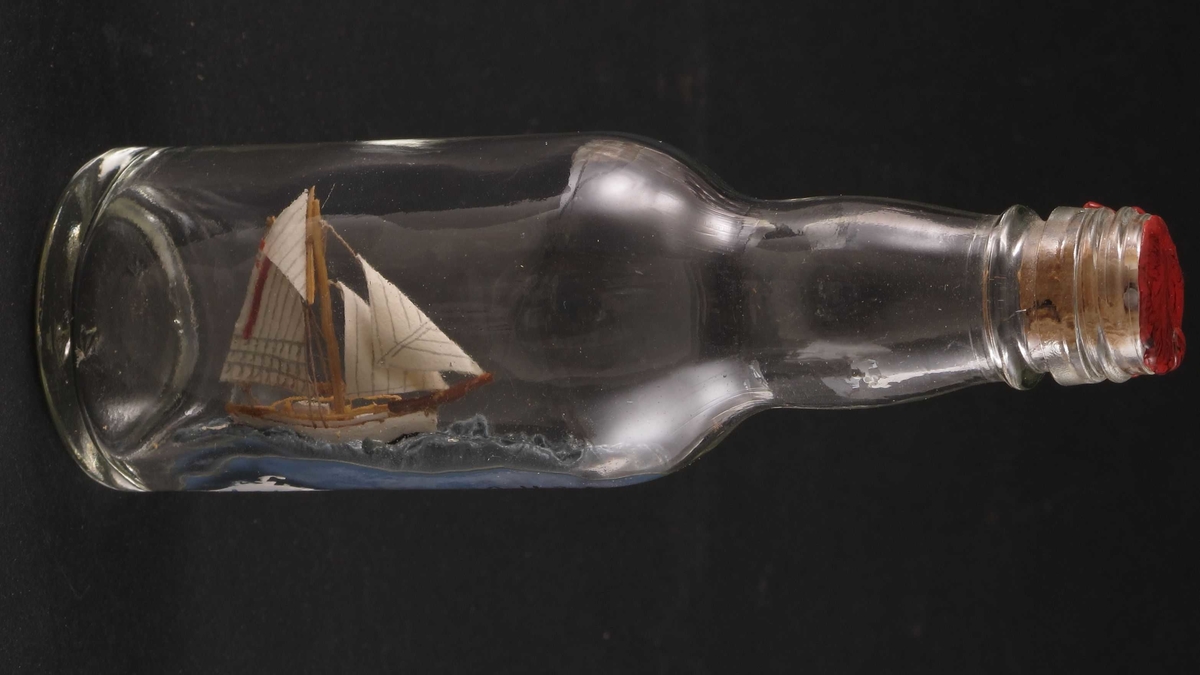 Mini flaskeskip: Losbåt.  Glass, klart. Kork med rød lakk. Hvitmalt miniatyrskute med rød bunn og rød stripe i seilet. Blå sjø.