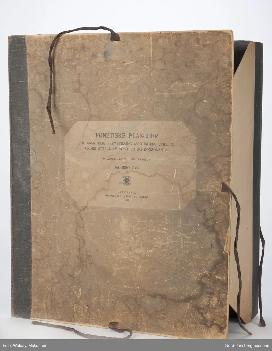 Pappomslag med knytebånd. Inneholder åtte plansjer som viser tungens stilling ved uttale av konsonater og vokaler.
Alle plansjene har opphengssnorer i tvunnet snor i rødt-hvitt-blått.