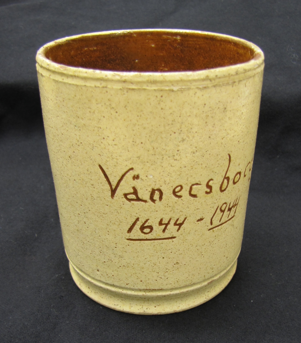 Keramikmugg med hänkel. Cylindrisk. Av keramik. Gul glasyr utvändigt med skriften "Vänersborg 1644 - 1944". Invändigt, brun glasyr.