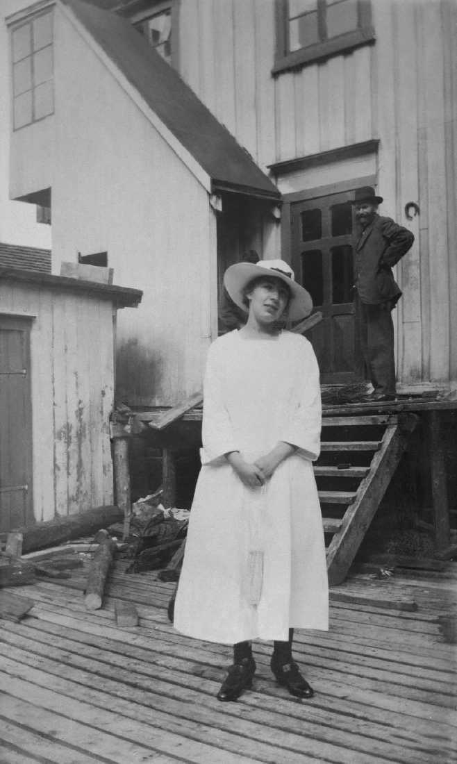 Kvinne i sommerkjole og hatt på baksiden av Barth`s butikk i Sjøgata 15