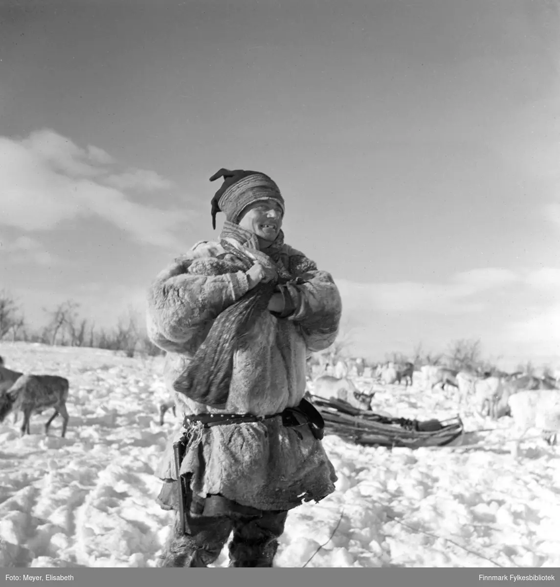 Henrik Andersen Spein kledd i pesk, fotografert på vidda i Kautokeino vinterstid. I bakgrunnen en slede og reinflokken.