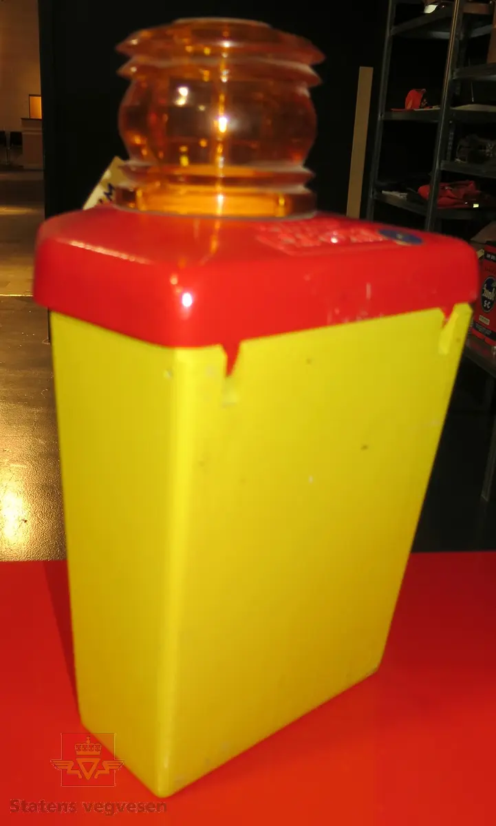 Varsellampe, blinkende type, blinker i alle retninger. Lagd av metall, i fargene gul og rød. Har en oransje plastkuppel på toppen. Drives av et 6 volt batteri (Airam 4R 20-7), dette mangler. Lampen kan startes og stoppes når du åpner lampehuset.