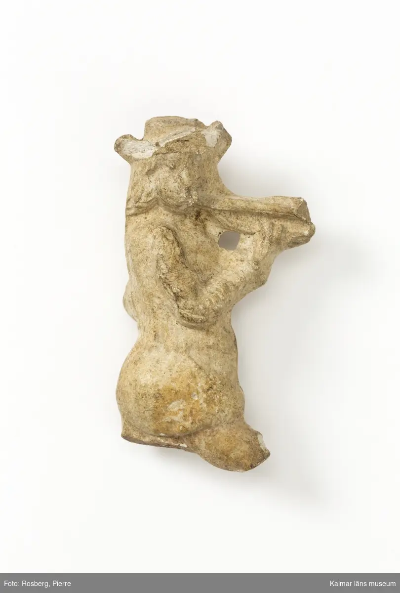 KLM 4070. Figur, av lera. Vit lera. Figuren visar en flöjt- eller lurblåsare i långt hår, hatt och kort jacka samt korta puffiga byxor. Tydliga gjutsömmar. Skadad, benen saknas. Datering, möjligen 1600-tal.